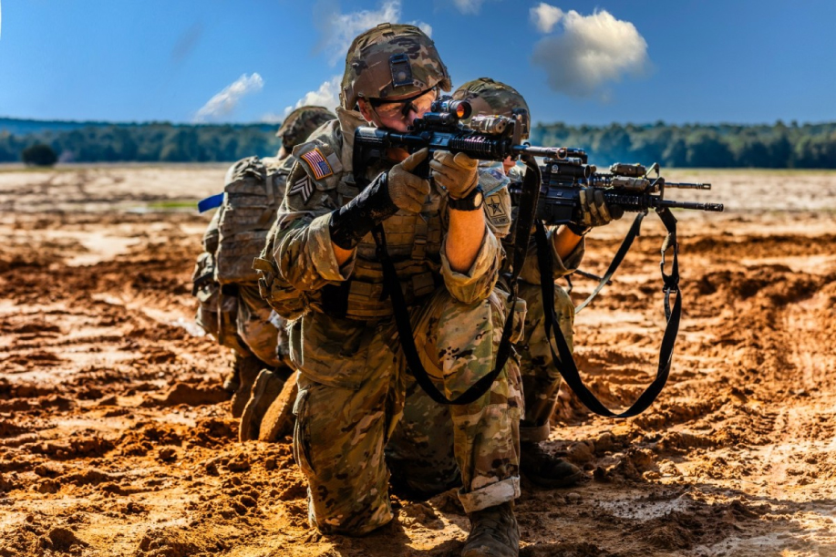 Three soldiers wearing combat gear aim weapons in different directions in a field during the day.