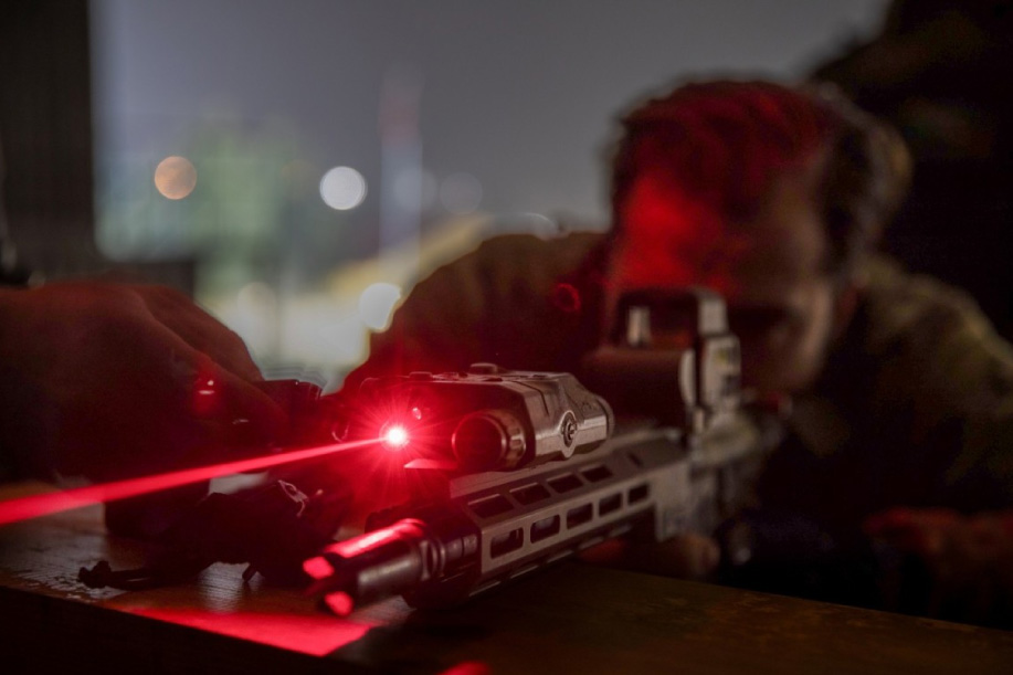 A soldier aims a weapon, which shoots out a red laser beam in a darkened area.