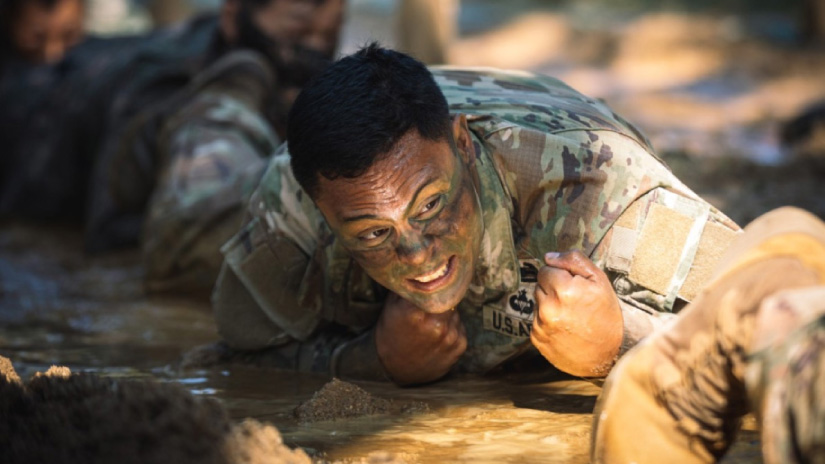 A soldier crawls under barbed wire while another soldier follows crawling close behind.