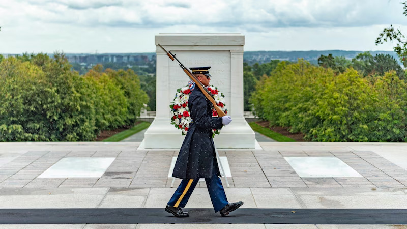 A soldier walks on a mat in front of a marble tomb.