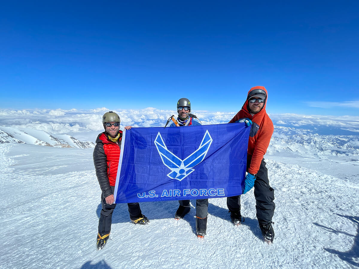 three men posing with air force flag on top of mountain